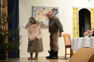 Theaterszene: Älteres Paar auf Bühne, Frau inspiziert Kleidungsstück, weihnachtliche Dekoration