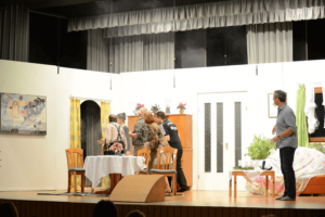 Mehrere Schauspieler auf Bühne verlassem eine Wohnzimmer-Kulisse, während ein Mann stehen bleibt