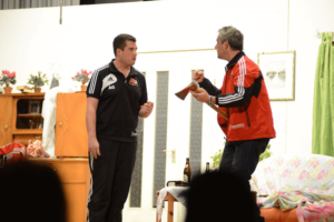 Zwei Darsteller sprechen miteinander mit Sportjacken und einer Axt