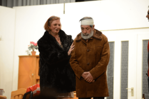 Zwei Darsteller, Mann und Frau nebeneinander, während der Mann mit Kopfbinde nach unten blickt und die Frau ihren Arm an ihn legt
