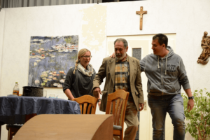 Drei Personen in einem Raum mit einem Jesuskreuz, während die mittlere Person umarmt wird