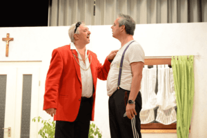 Theaterszene: Mann in rotem Jackett und Mann mit Hosenträgern in konfrontativem Dialog