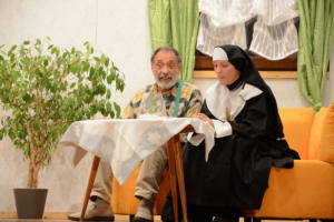 Theaterszene: Älterer Mann und Nonne an Tisch sitzend, Zimmerpflanze als Dekoration