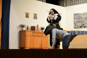Nonne auf Stuhl kämpfend mit Person am Boden, Heiligenbilder im Hintergrund