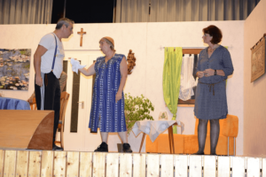 Theateraufführung mit drei Darstellern auf Bühne, Kreuz und Gemälde im Hintergrund