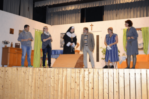 Theaterszene mit sechs Personen und Nonne auf Holzbühne, Kreuz an weißer Wand