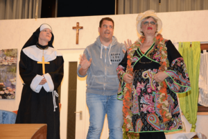 Theaterszene: Nonne, Mann in Kapuzenpulli und Person im bunten Kimono auf Kirchenbühne