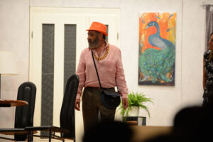 Mann in rosa Hemd mit orangem Hut und Goldkette vor Kunstgemälde